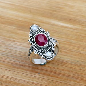 May include: A silver ring with a red gemstone surrounded by three white pearls. The ring has an ornate design.