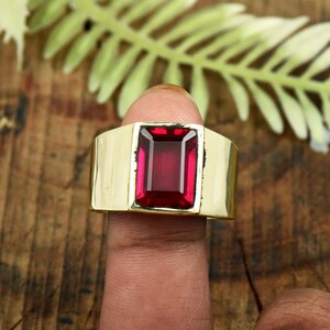 May include: A gold-toned ring featuring a large, rectangular, deep red gemstone set in the center. The band is wide and smooth with a polished finish, showcasing a bold design that highlights the vibrant color of the central stone.