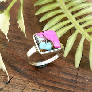 May include: A silver ring featuring a square-cut gemstone with a vibrant pink top, turquoise accents, and a silver bezel. The ring is set against a natural wood grain background, showcasing the jewelry's design and color.