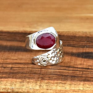 Puede incluir: Un anillo de plata con una piedra preciosa roja engastada en una banda martillada. El anillo está diseñado para envolverse alrededor del dedo.