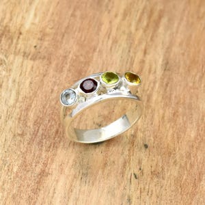 Peut inclure: Une bague en argent ornée de cinq pierres précieuses colorées : bleu clair, rouge foncé, vert clair et jaune. Les pierres sont serties sur un anneau argenté incurvé. La bague est présentée sur une surface en bois.