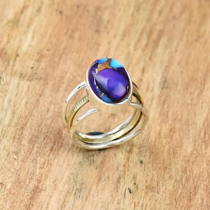 May include: A silver ring with a large, oval-shaped purple gemstone with blue and gold accents. The ring has a triple-band design with gold-colored accents on the bands.