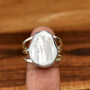 Puede incluir: Un anillo de plata con una perla grande, blanca e irregular en el centro. El anillo tiene un diseño de triple banda.