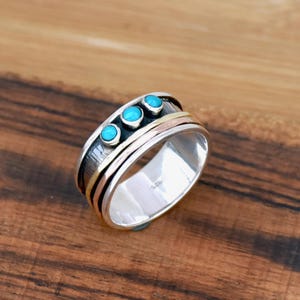 Peut inclure: Bague en argent ornée de trois pierres turquoise. La bague présente un design multicouche avec des bandes argentées, dorées et cuivrées. Posée sur une surface en bois.