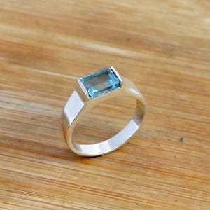 Op de afbeelding: Een zilveren ring met een lichtblauwe rechthoekige edelsteen.