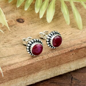 May include: A pair of sterling silver stud earrings with a round, deep red gemstone center. The earrings have a decorative, beaded border. The earrings are displayed on a wooden surface with green foliage in the background.