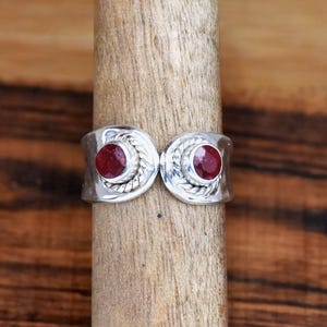 May include: A silver ring with two red gemstones set in the center of each side. The ring has a braided design around the gemstones.