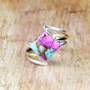May include: A silver ring with a square, multi-colored stone. The stone features pink, turquoise, and gold hues. The ring has a unique design with multiple bands, some of which are gold-toned. The ring is set against a wooden background.