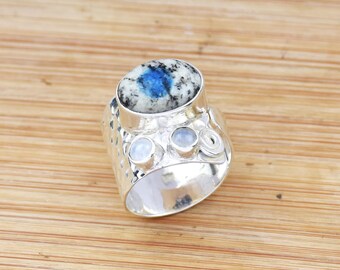 Könnte beinhalten: Ein silberner Ring mit einem großen ovalen K2-Stein mit blauen und weißen Mustern. Der Ring hat zwei kleinere runde Steine und ein strukturiertes Band. Der Ring liegt auf einer Holzoberfläche.