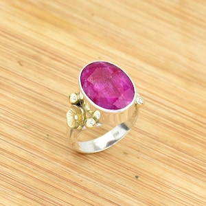 Peut inclure: Une bague en argent avec une grosse pierre précieuse magenta de coupe ovale. La bague présente des accents floraux dorés sur le bandeau. La pierre précieuse est le point central, sertie dans un bandeau argenté. La bague est présentée sur une surface en bois.