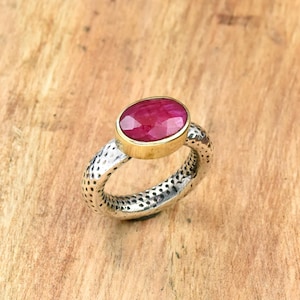Könnte beinhalten: Ein silberner Ring mit strukturiertem Band und einer goldenen Lünette, die einen großen, oval geschliffenen Rubin-Edelstein hält. Der Rubin hat eine tiefe, leuchtende rote Farbe, und der Ring ist vor einem Holzgrund platziert.