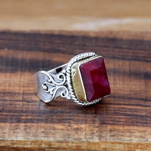 Könnte beinhalten: Ein silberner Ring mit einem roten Edelstein, der in einer goldenen Fassung sitzt. Der Ring hat ein dekoratives Band mit einem gedrehten Seilmuster.