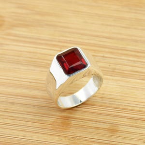 May include: A silver ring with a large, square, deep red gemstone. The ring has a wide band and a polished finish. The gemstone is set in a bezel setting, and the ring is displayed on a wooden surface.