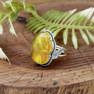 May include: A silver ring with a large, oval-shaped yellow gemstone. The gemstone has a swirling, translucent appearance. The ring features a detailed silver setting with a leaf design on the side. The ring is set against a wooden background.