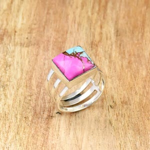 May include: A silver ring featuring a square-cut, vibrant pink stone with turquoise and brown accents. The ring has a multi-band design, with three parallel bands on each side of the stone, creating a modern and eye-catching look.