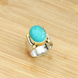 Puede incluir: Un anillo de plata con una piedra turquesa ovalada grande engastada en un bisel dorado. El anillo tiene una banda ancha con detalles dorados. Se muestra sobre una superficie de madera marrón claro.