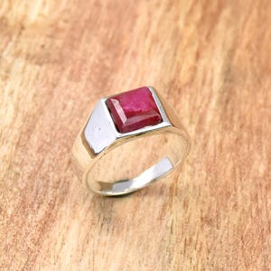 May include: A silver ring featuring a square, deep red gemstone. The ring has a simple, modern design with a polished finish. The gemstone is set in a bezel setting, and the band is smooth and wide.