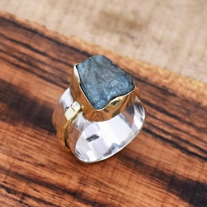 Könnte beinhalten: Ein Silber- und Goldring mit einem großen, rohen Aquamarin-Edelstein. Der Ring ist in einer breiten, strukturierten Fassung gefasst.