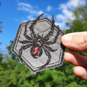 May include: A black embroidered spider patch with a red hourglass on a gray background. The patch is shaped like a hexagon and has a black stitched border.