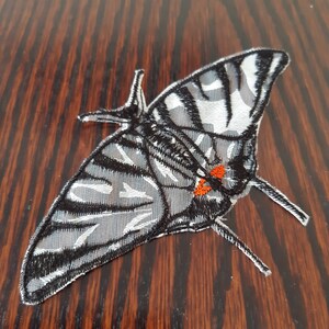 Può includere: Una toppa ricamata a forma di farfalla in bianco e nero con accenti arancioni. La farfalla è su una superficie di legno marrone.