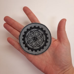 May include: A gray fabric patch with a black embroidered design. The design is a circular pattern with a central circle and four curved lines extending outward.