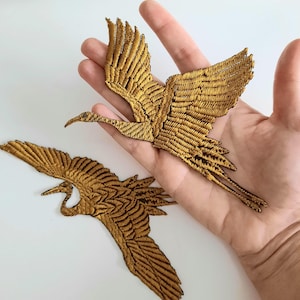 May include: Two gold embroidered crane patches. The cranes are in flight with their wings spread wide. The patches are ideal for adding a touch of elegance to clothing, bags, or other crafts.