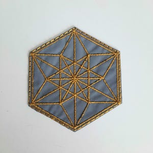 Puede incluir: Un posavasos hexagonal gris con un diseño geométrico dorado bordado en la superficie.