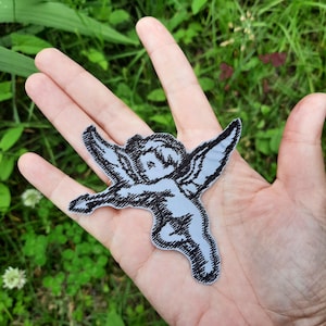 Pode incluir: Um remendo bordado em preto e branco de um querubim com asas, segurando um coração em suas mãos.