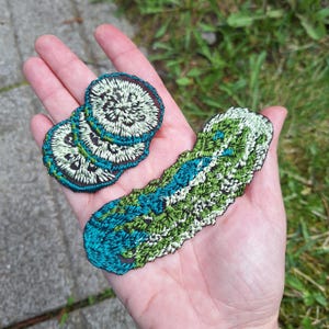 Może przedstawiać: Haftowane naszywki w odcieniach zieleni, turkusu i bieli. Jedna ma długi, zakrzywiony kształt, druga to stos trzech okrągłych naszywek. Naszywki trzymane są w dłoni na tle zielonej trawy i szarej powierzchni.