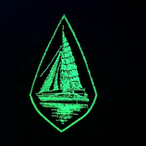 Könnte beinhalten: Ein grüner, im Dunkeln leuchtender gestickter Aufnäher in Tropfenform mit einem Segelboot-Motiv im Inneren.