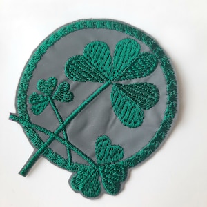 Puede incluir: Un parche bordado de trébol verde sobre un fondo gris con un contorno blanco. El trébol es un símbolo de buena suerte y a menudo se asocia con Irlanda.