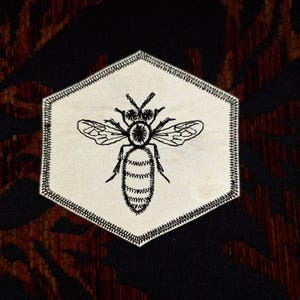 May include: A black and white embroidered bee patch in a hexagonal shape. The bee is centered on a cream-colored background with a black stitched border. The background is a mix of black and brown.