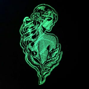 May include: A green glow-in-the-dark embroidered patch featuring a woman with long hair and a floral crown. The woman is facing to the right and her body is turned slightly to the left. The patch is made of a soft, textured fabric.