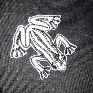 以下が含まれることがあります： 黒い布地に白い刺繍の蛙のワッペン