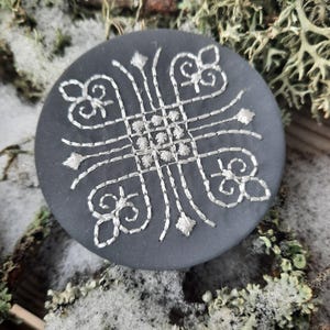 May include: A round, black felt pin with a white embroidered design. The design features a geometric pattern with a central square and four curved lines extending from each corner.