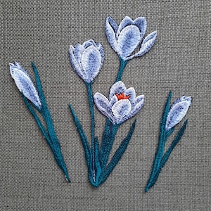 Könnte beinhalten: Gesticktes Blumendesign mit blau-weißen Krokusblüten mit orangefarbenen Zentren und grünen Stielen und Blättern. Das Design ist vor einem strukturierten grauen Hintergrund platziert und zeigt die detaillierten Nähte und den Farbkontrast.