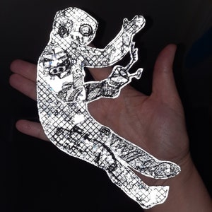 Könnte beinhalten: Ein reflektierender silberner Aufkleber mit einer stilisierten Figur in einer dynamischen Pose. Die Figur ist schwarz umrandet und hat ein Rastermuster über die gesamte Fläche.