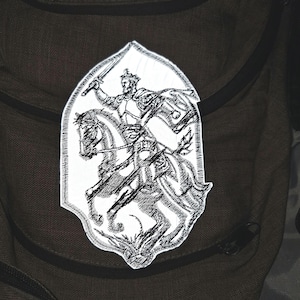 May include: A white embroidered patch featuring a black line art illustration of a knight in armor wearing a crown and riding a horse while holding a sword aloft. The patch has a pointed, shield-like shape with a stitched border edge.