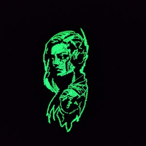 Puede incluir: Un diseño bordado en verde neón sobre un fondo negro. El diseño representa el perfil de una persona, con auriculares y un tatuaje detallado en el hombro. El estilo de bordado da un efecto texturizado y brillante.