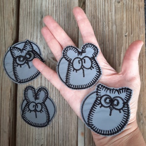 May include: Four gray felt patches with black embroidered cartoon cat faces. The cat faces have large eyes and a simple smile.