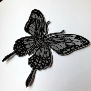 May include: Black and grey embroidered butterfly patch. The butterfly has detailed wings and a textured surface.