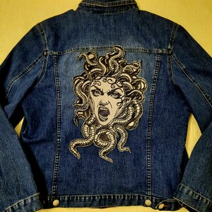 Op de afbeelding: Een blauwe denimjas met een zwart-witte geborduurde patch van Medusa, een mythische Griekse figuur met slangenhaar, op de rug.