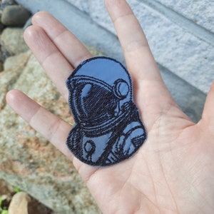 Puede incluir: Un parche bordado azul de un astronauta que lleva un casco con una visera negra. El astronauta está mirando hacia el lado derecho de la imagen.