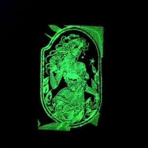 Może przedstawiać: Zielona, świecąca w ciemności, haftowana naszywka przedstawiająca kobietę z długimi włosami i zwiewną suknią. Naszywka jest otoczona ozdobną ramką.
