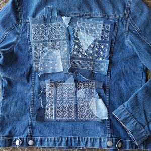 Könnte beinhalten: Eine blaue Jeansjacke mit weißen Stickereien, die auf den Rücken genäht sind. Die Patches haben verschiedene geometrische Muster.