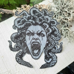 Puede incluir: Un parche bordado en blanco y negro de Medusa, una figura mitológica griega con serpientes por cabello, con una expresión feroz en su rostro.