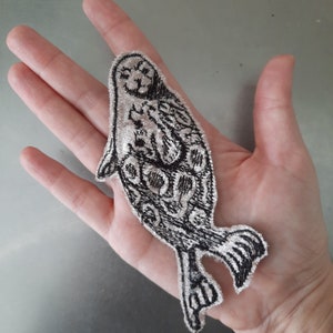 May include: A black and white embroidered iron-on patch of a seal. The seal is curled up with its flippers tucked under its body. The patch is on a hand.