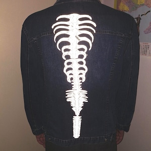 Könnte beinhalten: Eine dunkelblaue Jeansjacke mit einem großen, weißen, reflektierenden Grafikdruck eines menschlichen Brustkorbs und einer Wirbelsäule, die vertikal in der Mitte des Rückens verläuft. Die Jacke hat lange Ärmel, einen Kragen und sichtbare Nähte.