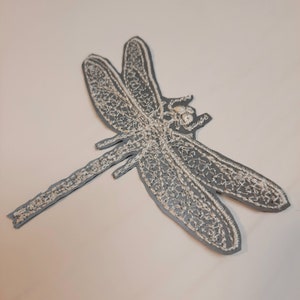Op de afbeelding: Een witte geborduurde libellenpatch op een grijze achtergrond. De libel is gedetailleerd met ingewikkelde steken en heeft een lang, dun lichaam.