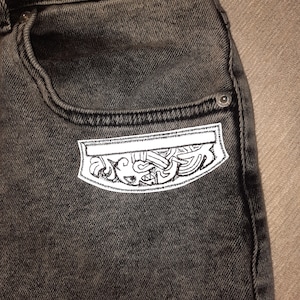 May include: A pair of black denim jeans with a white embroidered patch on the back pocket. The patch features a detailed design of a stylised creature.